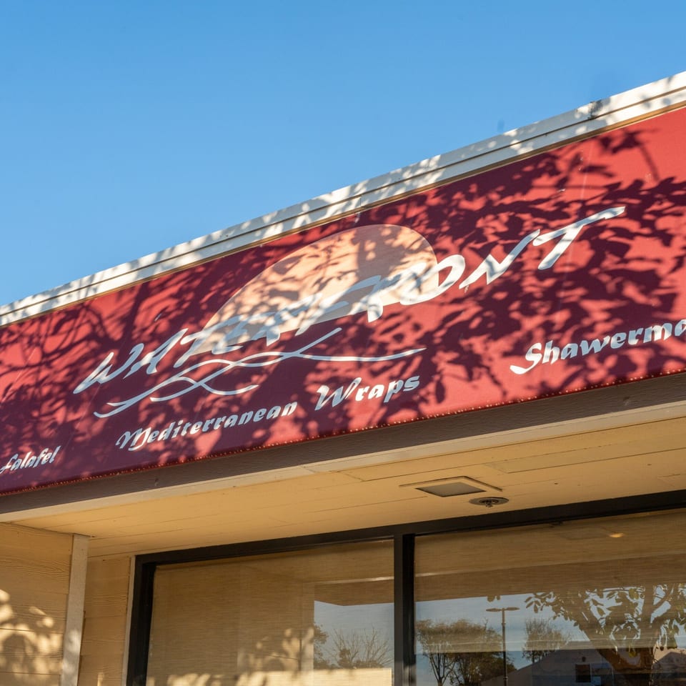 Red awning with "Waterfront Mediterranean Wraps" and "Shawerma" text, casting tree shadows.