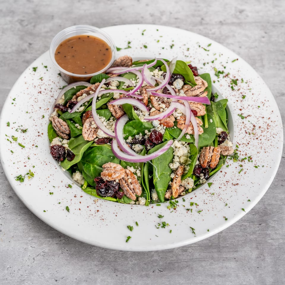 Spinach salad with red onions, pecans, dried cranberries, blue cheese, and a side of dressing.