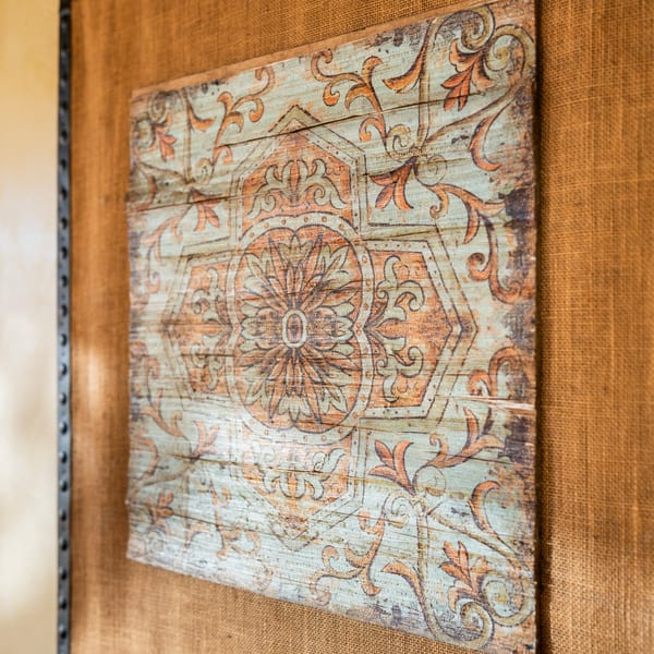 Distressed wooden wall art with an ornate orange and blue floral pattern on a burlap background.