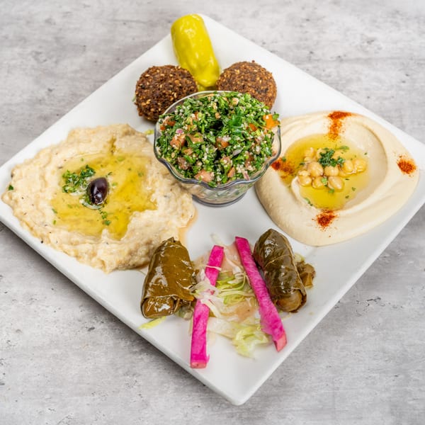 A white square plate filled with various Mediterranean appetizers, including hummus, baba ghanoush, falafel, tabbouleh, and stuffed grape leaves.