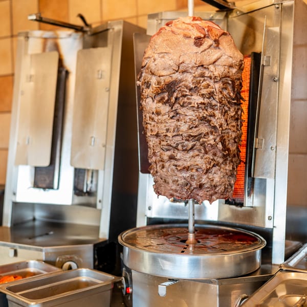 A large vertical spit of rotating meat cooking next to a fiery grill in a kitchen.